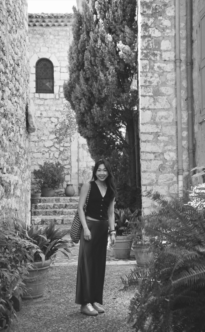 Ming in a black-and-white photo, smiling in a stone lane lined with potted plants and a cypress tree