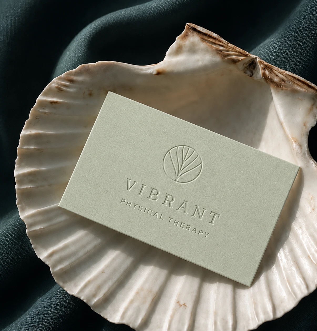 Sage business card with the blind-embossed Vibrant Physical Therapy logo, resting in a white scallop shell on deep teal linen